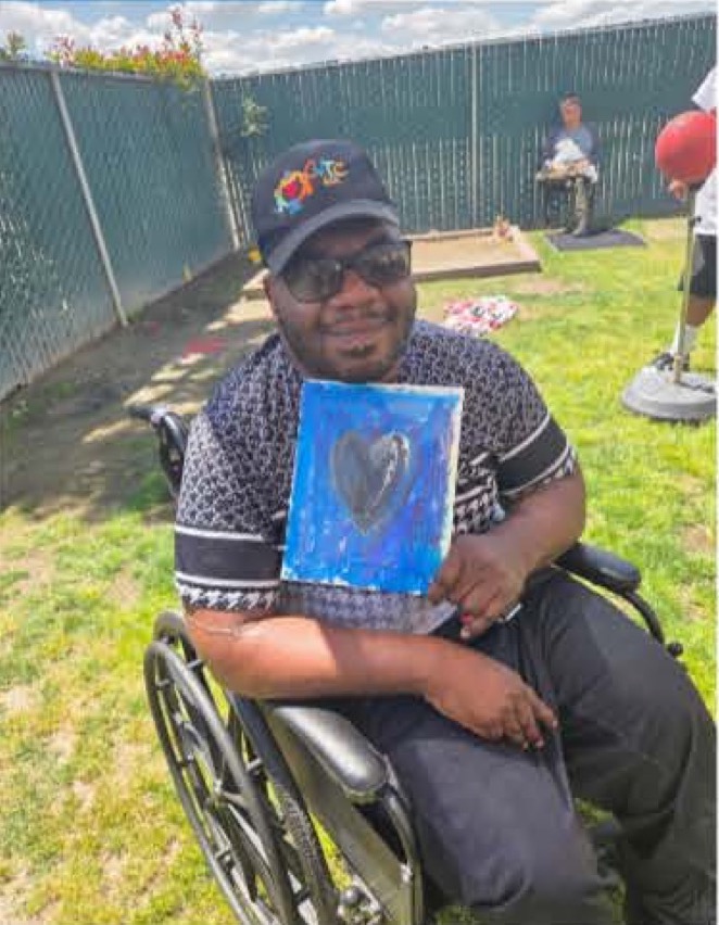 Alex B. smiling in his wheelchair while holding one of his paintings outdoors at the Visalia program.