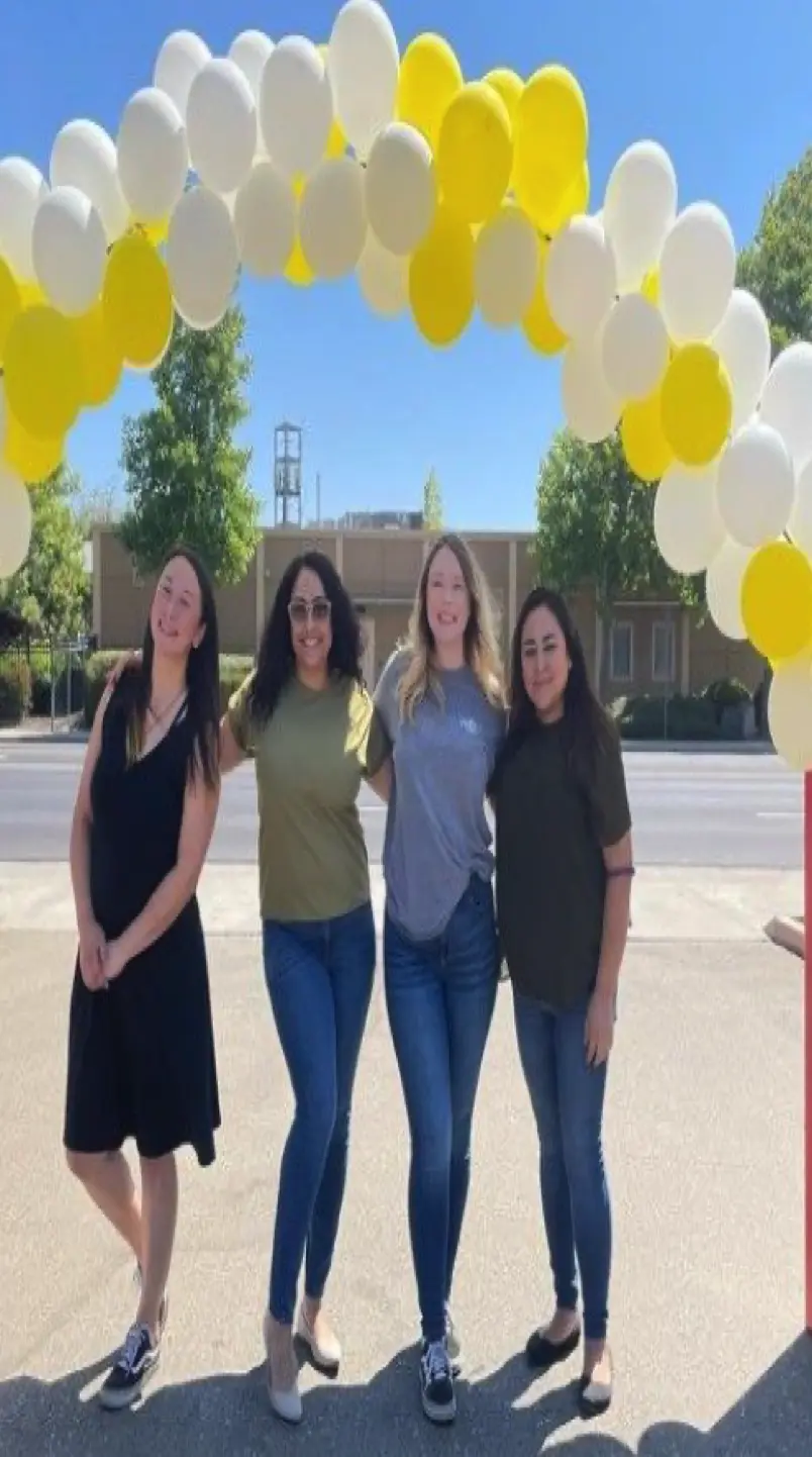 A group of smiling women standing under yellow and white balloons.